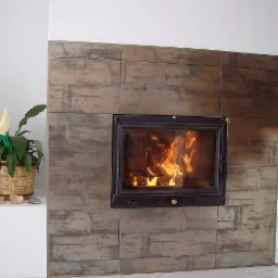 Obudowa kominka wyłożona płytkami imitującymi beton, z paleniskiem, w którym płonie ogień, obok stoi doniczka z kwiatem.