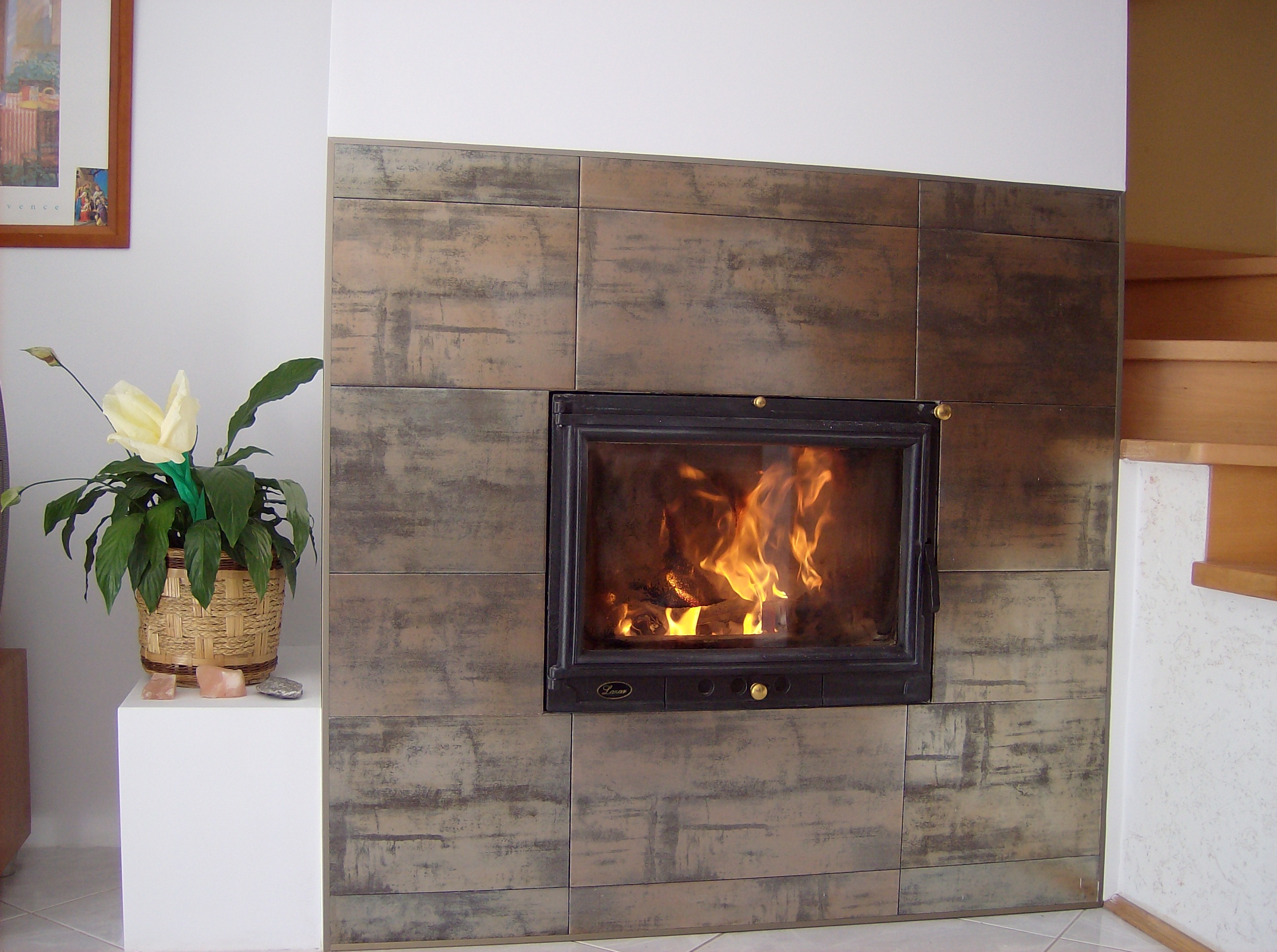 Obudowa kominka wyłożona płytkami imitującymi beton, z paleniskiem, w którym płonie ogień, obok stoi doniczka z kwiatem.