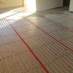 Rozłożone rury ogrzewania podłogowego na folii z czerwoną siatką, widok z góry, jasne światło słoneczne wpadające przez okno.