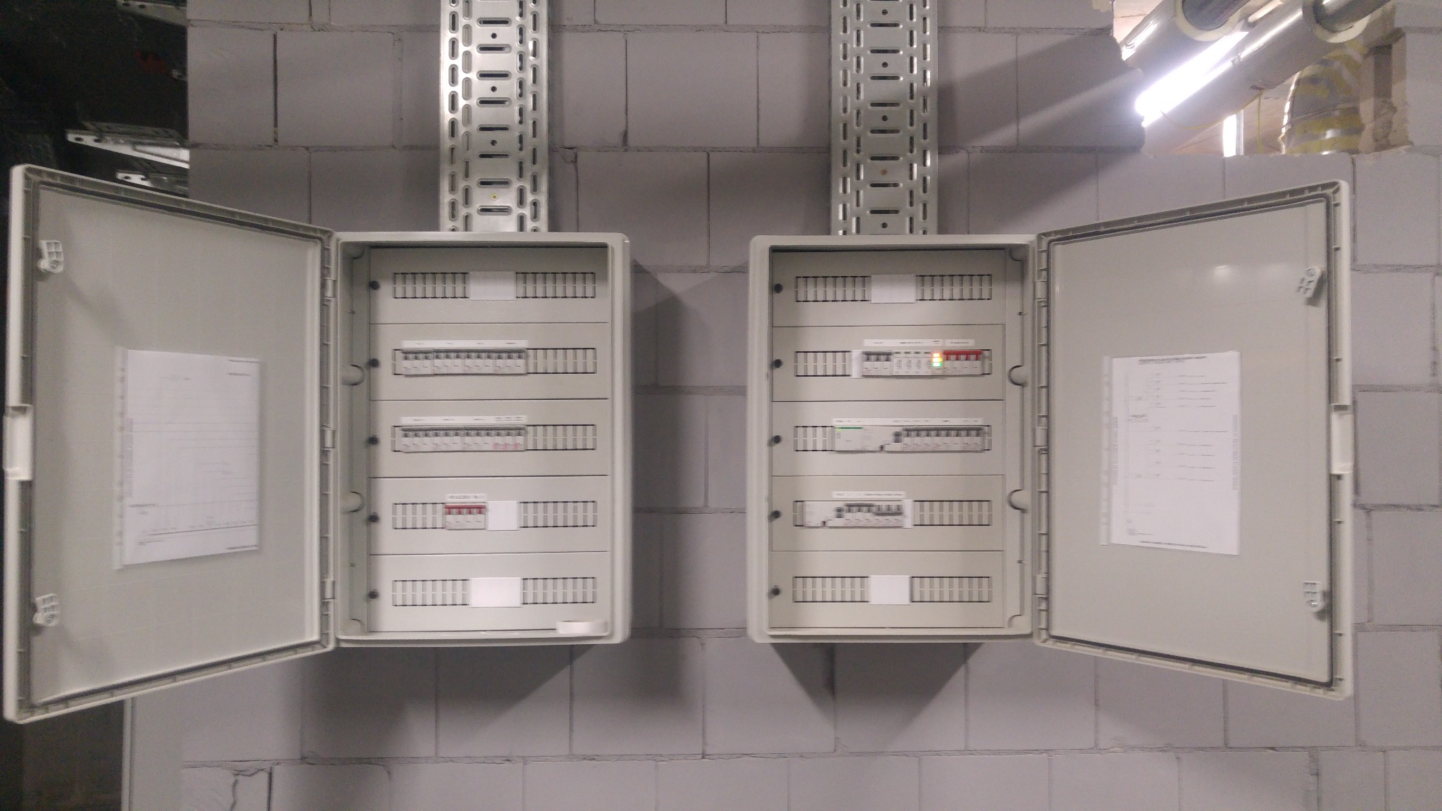 Dwa otwarte panele elektryczne na szarej ścianie z betonowych bloków, widoczne wyłączniki i dokumentacja wewnątrz, nad nimi metalowe korytka kablowe.