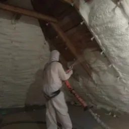 Pracownik w kombinezonie ochronnym aplikuje piankę izolacyjną na poddaszu w Mińsku Mazowieckim, widoczne drewniane elementy konstrukcji dachu i gruba warstwa nałożonej pianki.