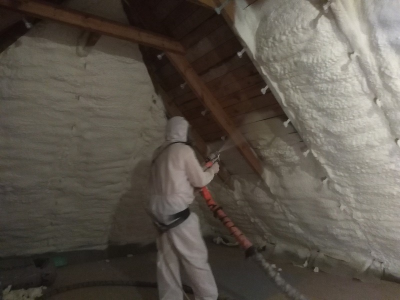 Pracownik w kombinezonie ochronnym aplikuje piankę izolacyjną na poddaszu w Mińsku Mazowieckim, widoczne drewniane elementy konstrukcji dachu i gruba warstwa nałożonej pianki.