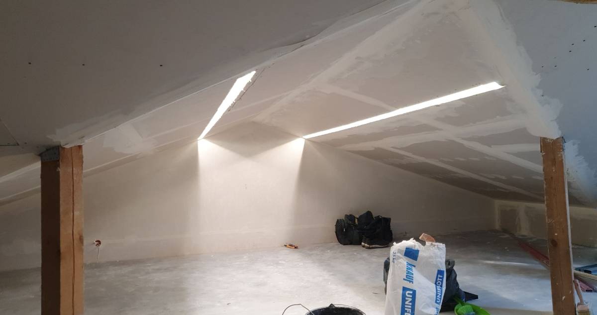 Poddasze z ukończonymi pracami montażu płyt kartonowo-gipsowych, widoczne łączenia szpachlowane, drewniane słupy nośne i worki z materiałami budowlanymi na podłodze.