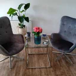 Dwa szare, welurowe fotele na chromowanych nogach, szklany stolik kawowy z bukietem róż i filiżanką w gabinecie.