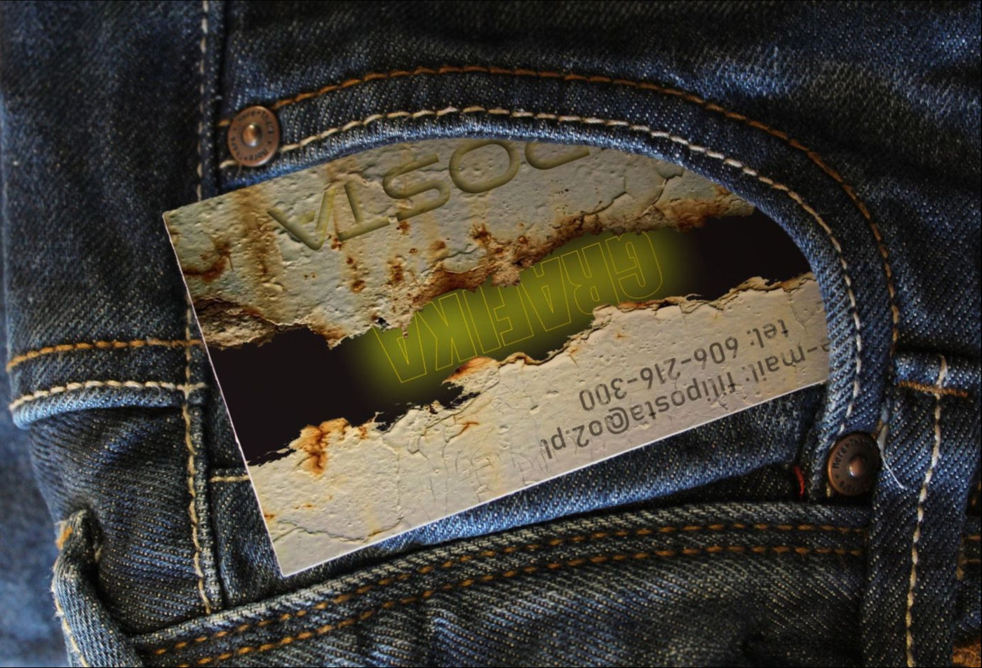 Wizytówka z efektem zniszczonej ściany i zielonym napisem 'GRAFIKA' wystająca z kieszeni dżinsów.