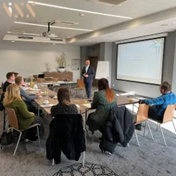 Prelegent w garniturze prowadzi prezentację dla grupy osób siedzących przy stole w sali konferencyjnej, na ekranie wyświetlony napis 'Czy jestem asertywny?'