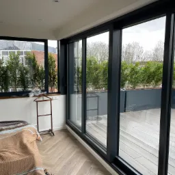 Nowoczesne okno przesuwne aluminiowe z widokiem na taras z drewnianą podłogą i roślinami, widoczne fragmenty sypialni z parkietem w jodełkę i łóżkiem.