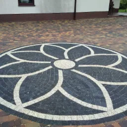 Okrągły wzór kwiatowy ułożony z czarnej i białej kostki brukowej na tle wielokolorowej kostki, widok z góry, obok budynek z ceglaną podmurówką.