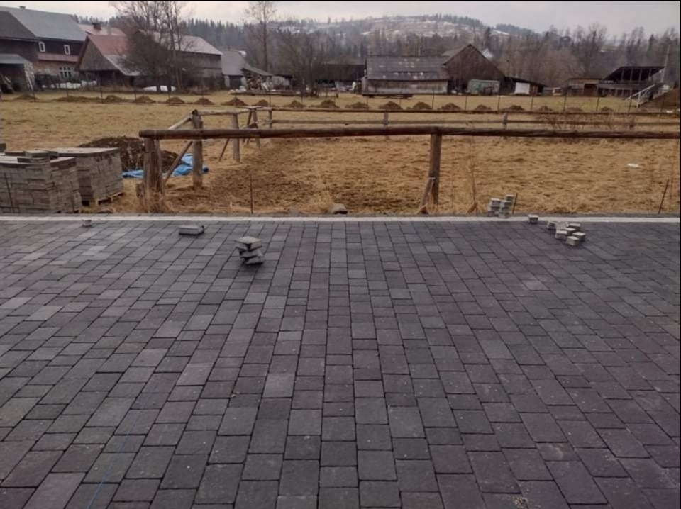 Częściowo ułożona, szara kostka brukowa na posesji wiejskiej, z widocznymi stertami kostki i drewnianym ogrodzeniem w tle.