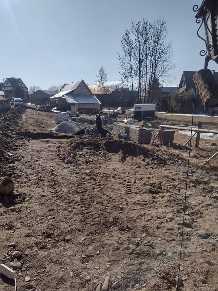 Przygotowanie terenu pod układanie kostki brukowej na tle górskiego krajobrazu z widocznym budynkiem w trakcie budowy.