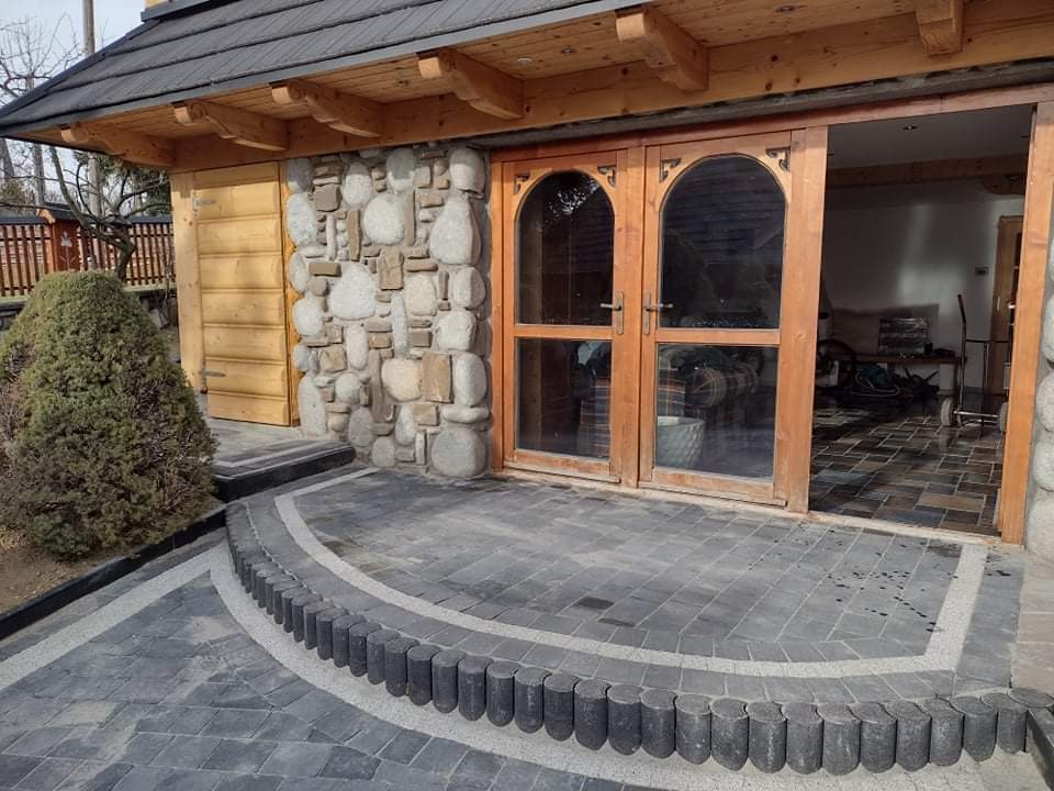 Ułożona kostka brukowa w kolorze grafitowym z obrzeżem z drewnianych palisad, prowadząca do kamiennej ściany z drewnianymi drzwiami łukowymi.