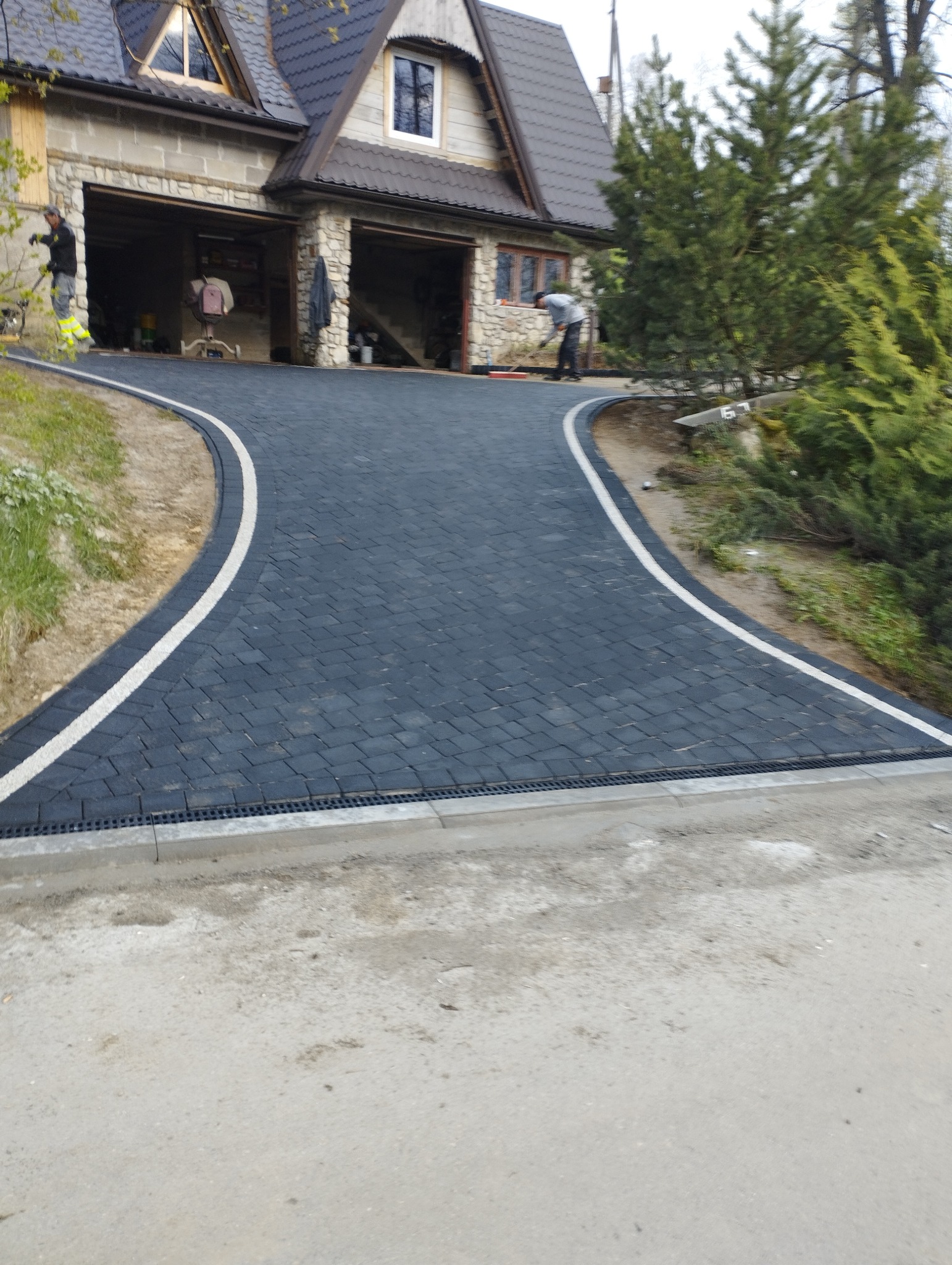 Czarna kostka brukowa ułożona na podjeździe do domu z dwoma garażami, wykończona białym obramowaniem i betonowym krawężnikiem z odpływem liniowym. Pracownicy w tle.