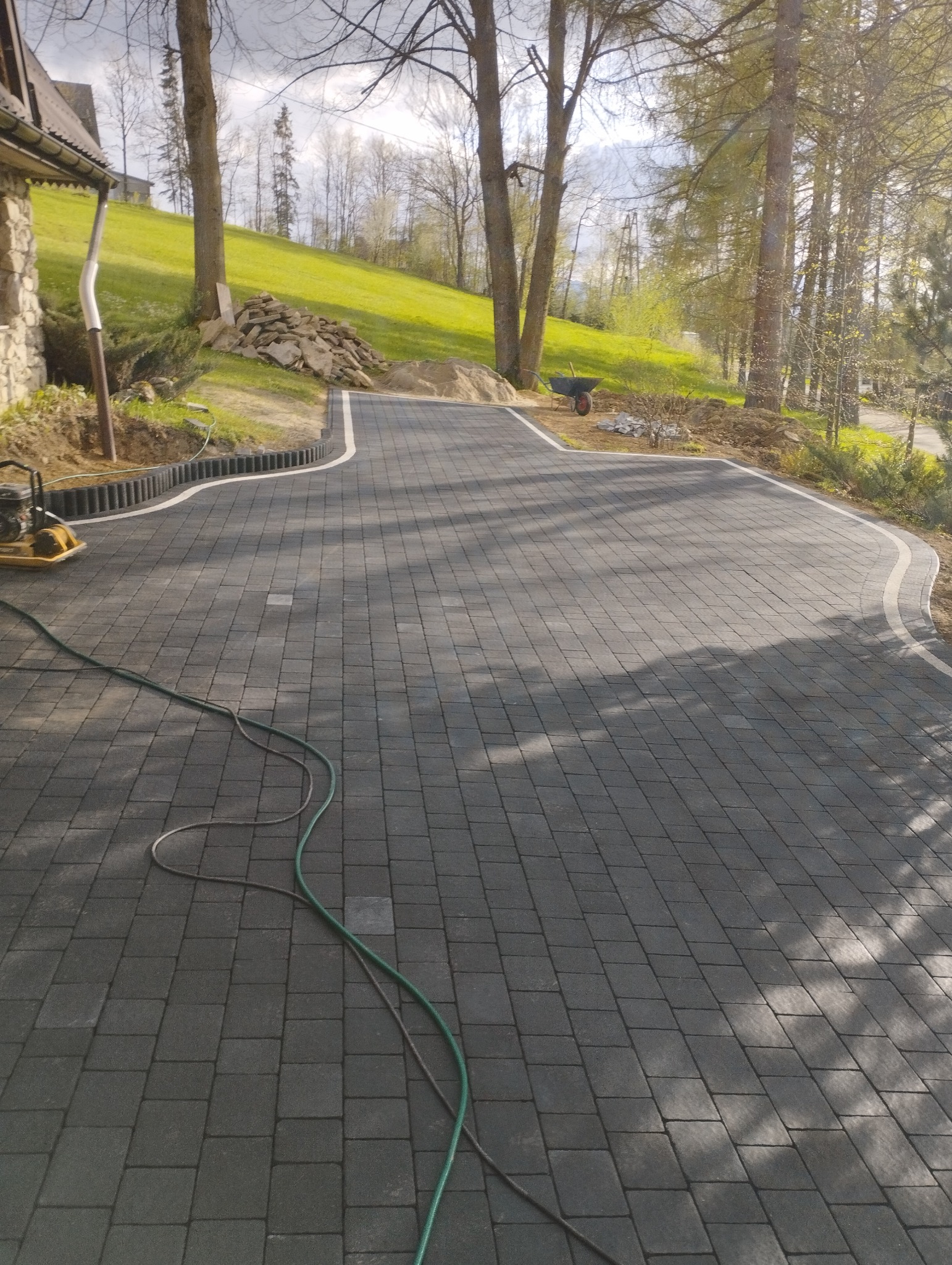 Ułożona, ciemnoszara kostka brukowa na podjeździe, z widocznym obrzeżem z elementami imitującymi drewno, sprzęt do zagęszczania i wąż na powierzchni, w tle trawnik na wzgórzu i drzewa.