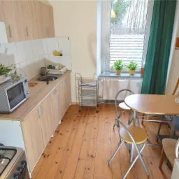 Jasna kuchnia z drewnianą podłogą, meblami w odcieniach beżu i stołem z krzesłami. Na blacie kuchennym mikrofalówka z doniczką z ziołami. Za oknem widok na drzewa.