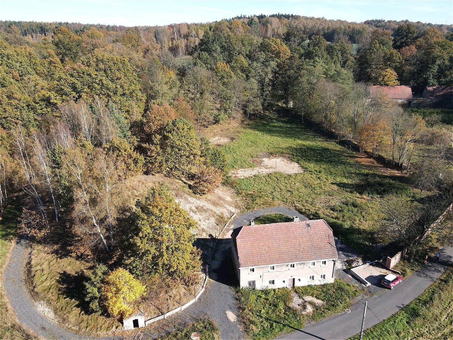 Widok z lotu ptaka na dwupiętrowy dom z czerwoną dachówką otoczony jesiennym lasem, z widocznym podjazdem i czerwonym samochodem obok fundamentów.