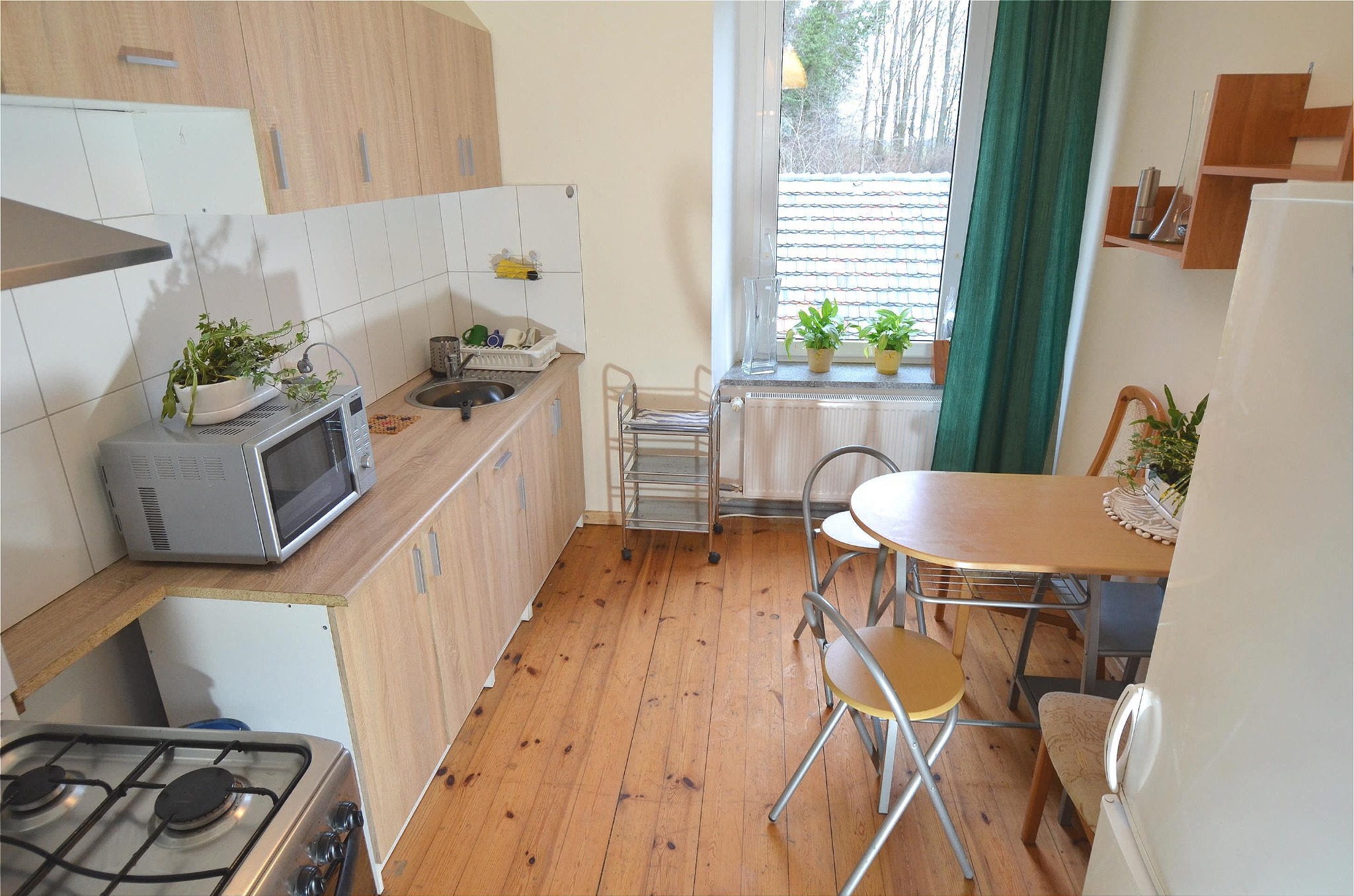 Jasna kuchnia z drewnianą podłogą, meblami w odcieniach beżu i stołem z krzesłami. Na blacie kuchennym mikrofalówka z doniczką z ziołami. Za oknem widok na drzewa.