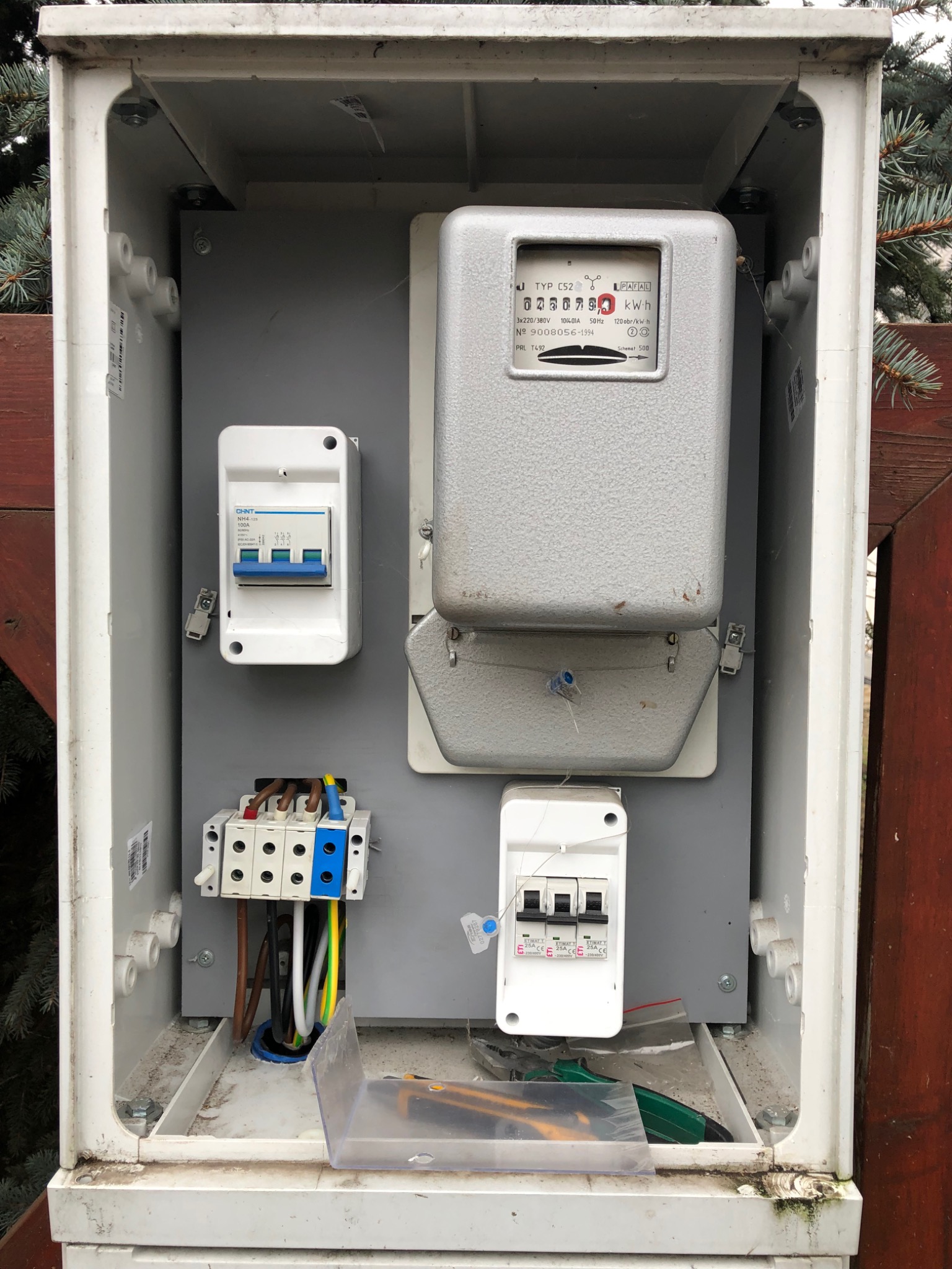 Otwarta skrzynka elektryczna z zamontowanym licznikiem energii, bezpiecznikami i złączkami przewodów, widoczne także narzędzia: nóż i szczypce.