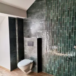 Nowoczesna łazienka z wiszącą toaletą, zielonymi, błyszczącymi płytkami wertykalnymi i drewnopodobną podłogą. Widoczne czarne elementy armatury.