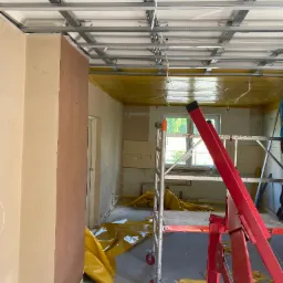 Montaż metalowego stelażu sufitu podwieszanego w trakcie remontu. Widoczne rusztowanie i folia ochronna. W tle okno z widokiem na zieleń.