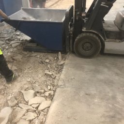 Dominik Micun - Praca w toku: operator wózka widłowego transportuje gruz w niebieskim kontenerze. W tle widoczny pracownik w ochronnikach słuchu. Podłoże częściowo oczyszczone z resztek.