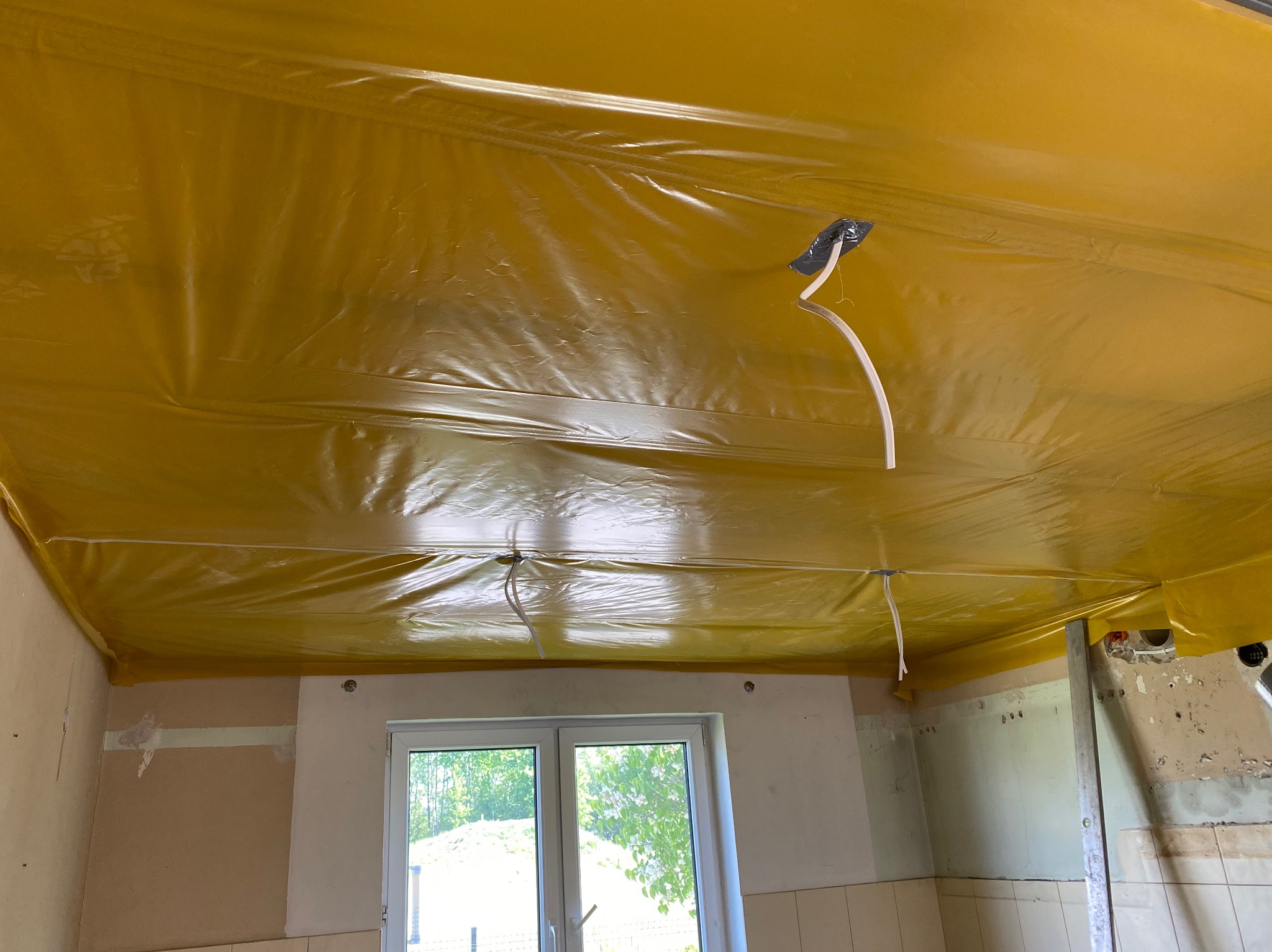 Żółta folia zabezpieczająca na suficie w trakcie remontu, widoczne kable elektryczne i fragment okna. Przygotowanie do prac wykończeniowych.
