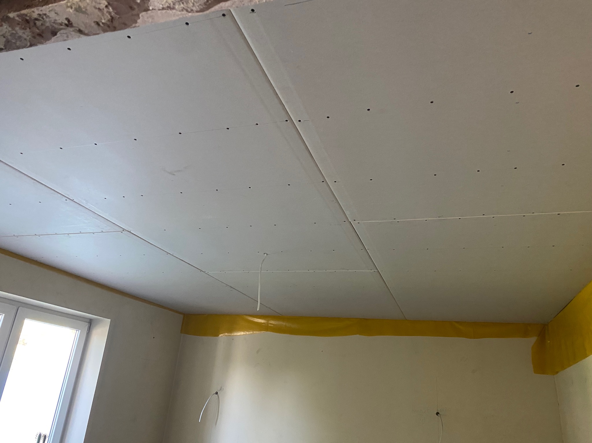 Zamontowany sufit z płyt kartonowo-gipsowych, widoczne łączenia i otwory na wkręty, zabezpieczone ściany taśmą malarską, okno z lewej strony kadru.