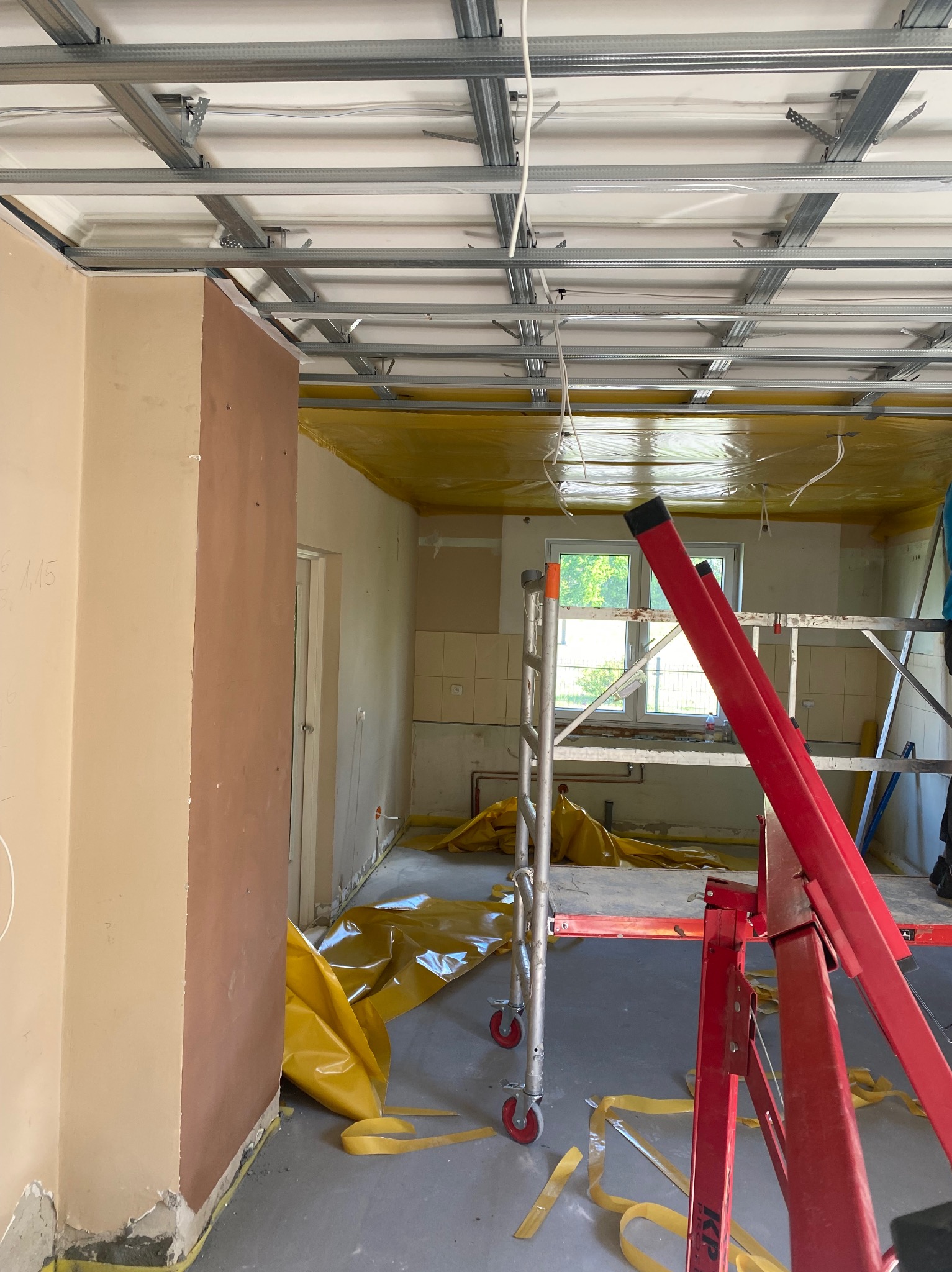 Montaż metalowego stelażu sufitu podwieszanego w trakcie remontu. Widoczne rusztowanie i folia ochronna. W tle okno z widokiem na zieleń.