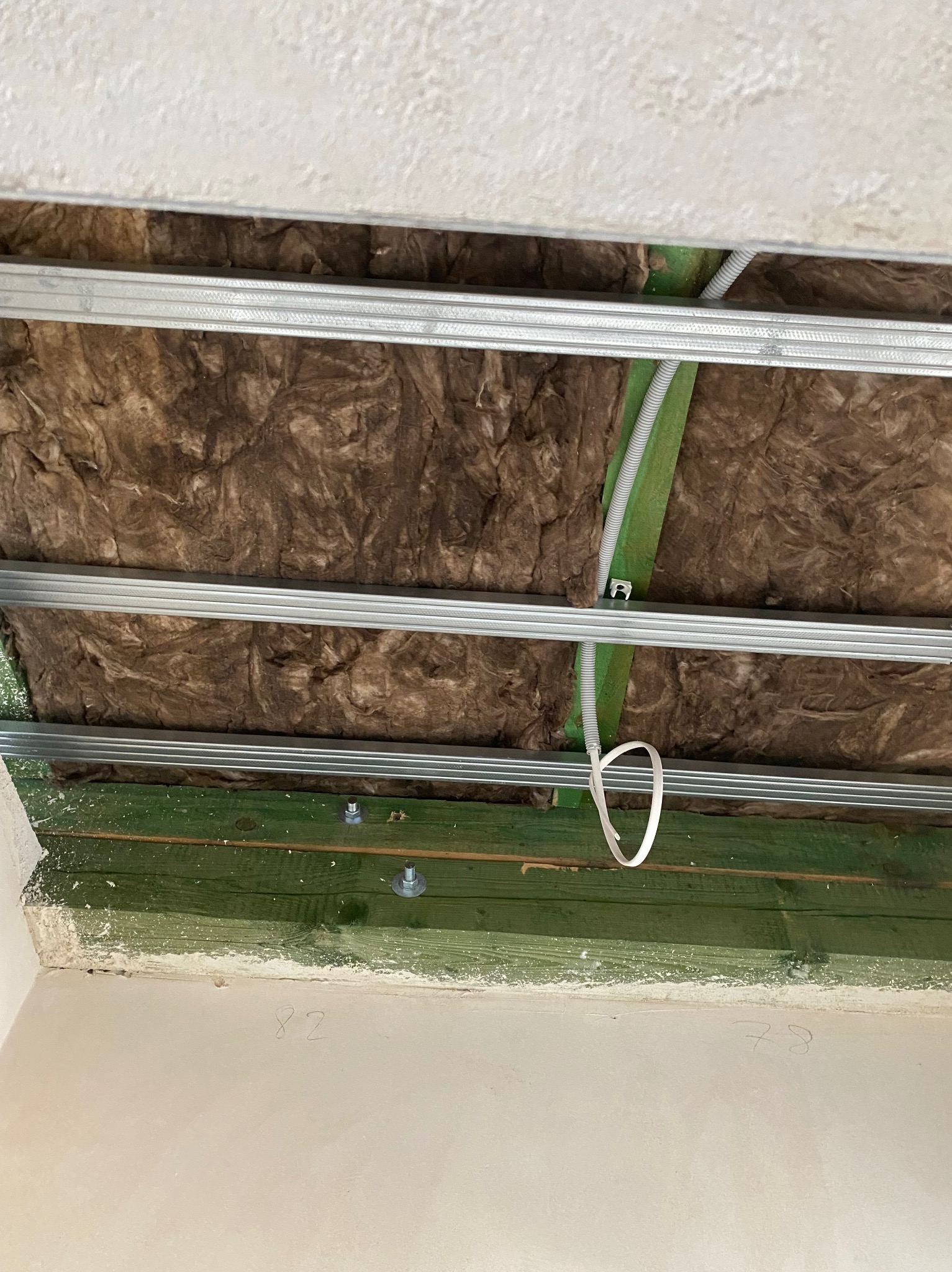 Poddasze w trakcie ocieplania: wełna mineralna między metalowymi profilami, widoczne zielone drewniane elementy konstrukcji i fragment instalacji elektrycznej.