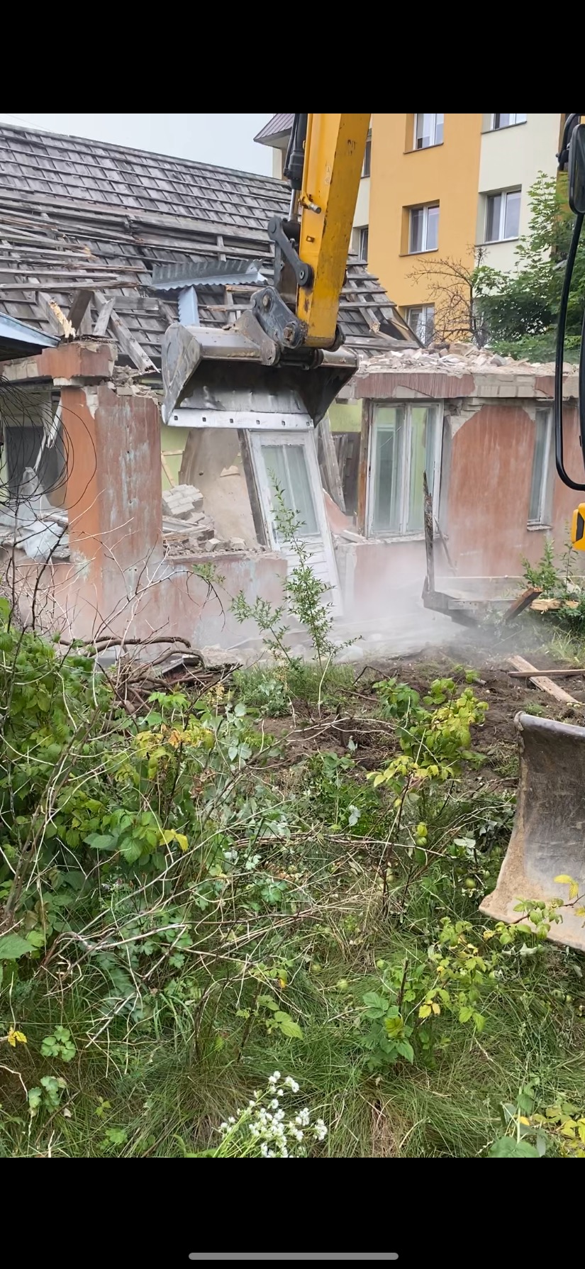 Żółta koparka z łyżką w trakcie wyburzania starego, ceglanego budynku z drewnianą stolarką okienną. Widoczne gruz i zieleń. Praca w toku.