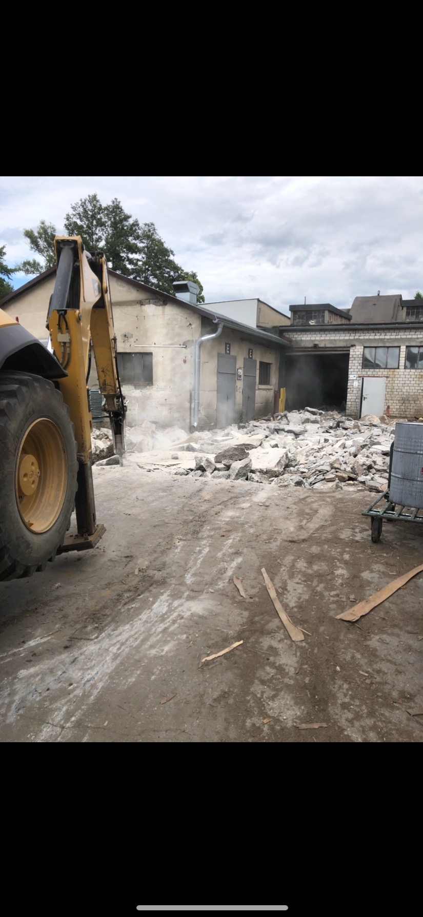 Żółta koparka z młotem pneumatycznym kruszy beton przed budynkiem magazynowym. Widoczne gruz i pył. Etap rozbiórki, dzień pochmurny.