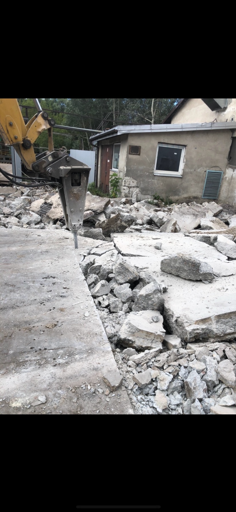 Żółty młot pneumatyczny kruszy betonową posadzkę przed budynkiem. Gruz i odpryski porozrzucane wokół. Widoczne elementy budynku w tle.