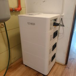 MelSol Mariusz Setlak - Biały, modułowy magazyn energii Dyness z podłączonymi kablami, ustawiony w rogu pomieszczenia z drewnopodobną podłogą i instalacjami na ścianach.