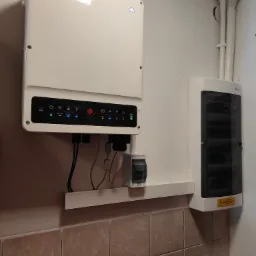 Biały inwerter fotowoltaiczny z diodami LED i obudową elektryczną zamontowany na ścianie z beżowymi płytkami. Widoczne przewody i wyłącznik.