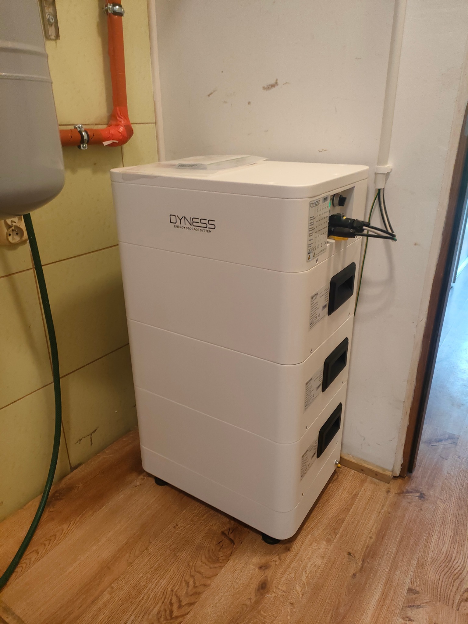 Biały, modułowy magazyn energii Dyness z podłączonymi kablami, ustawiony w rogu pomieszczenia z drewnopodobną podłogą i instalacjami na ścianach.