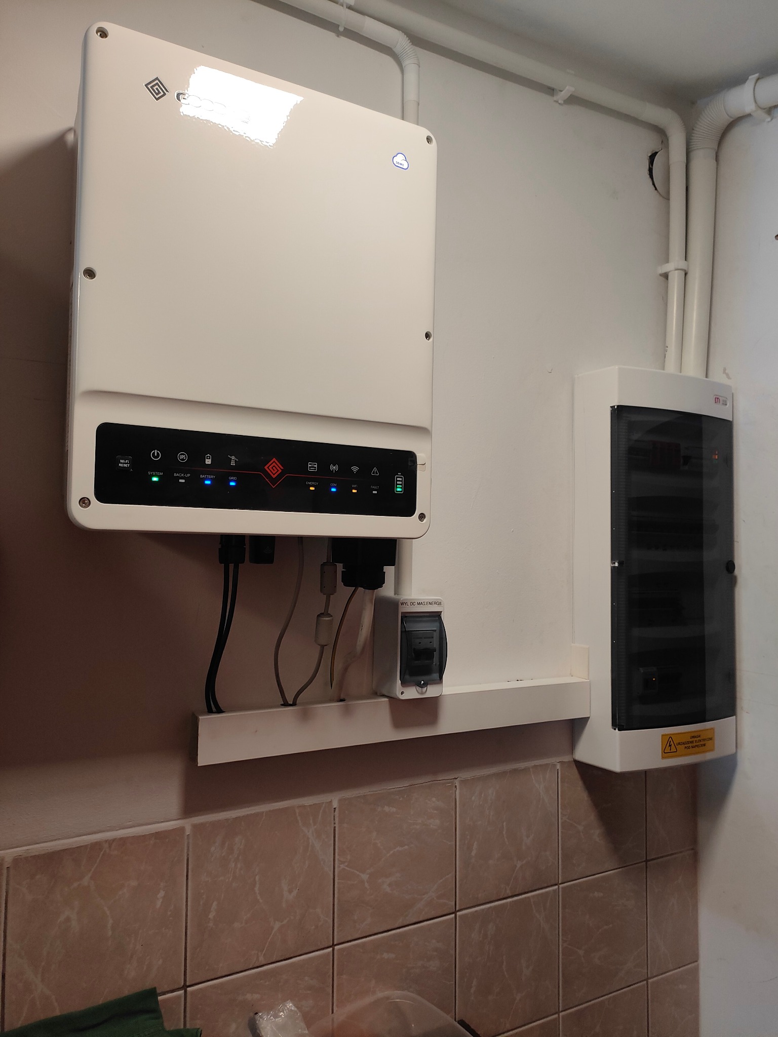 Biały inwerter fotowoltaiczny z diodami LED i obudową elektryczną zamontowany na ścianie z beżowymi płytkami. Widoczne przewody i wyłącznik.