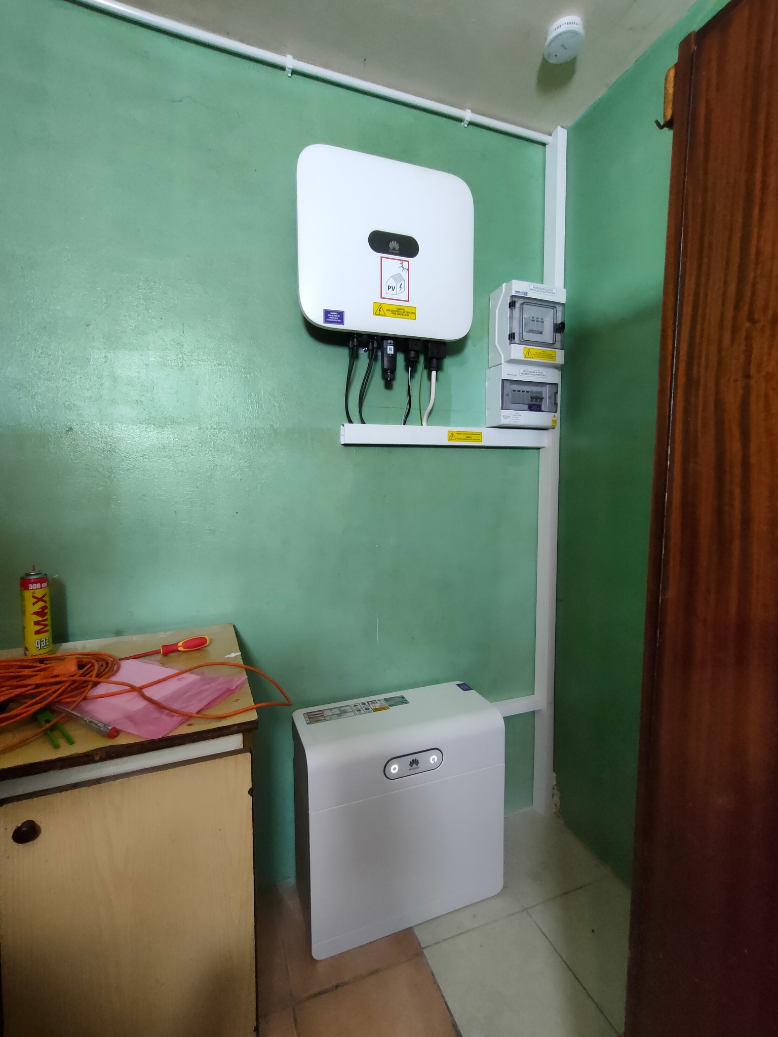 Biały inwerter fotowoltaiczny Huawei zamontowany na zielonej ścianie, obok rozdzielnica i akumulator. Na blacie narzędzia i przewody. Widok instalacji fotowoltaicznej.