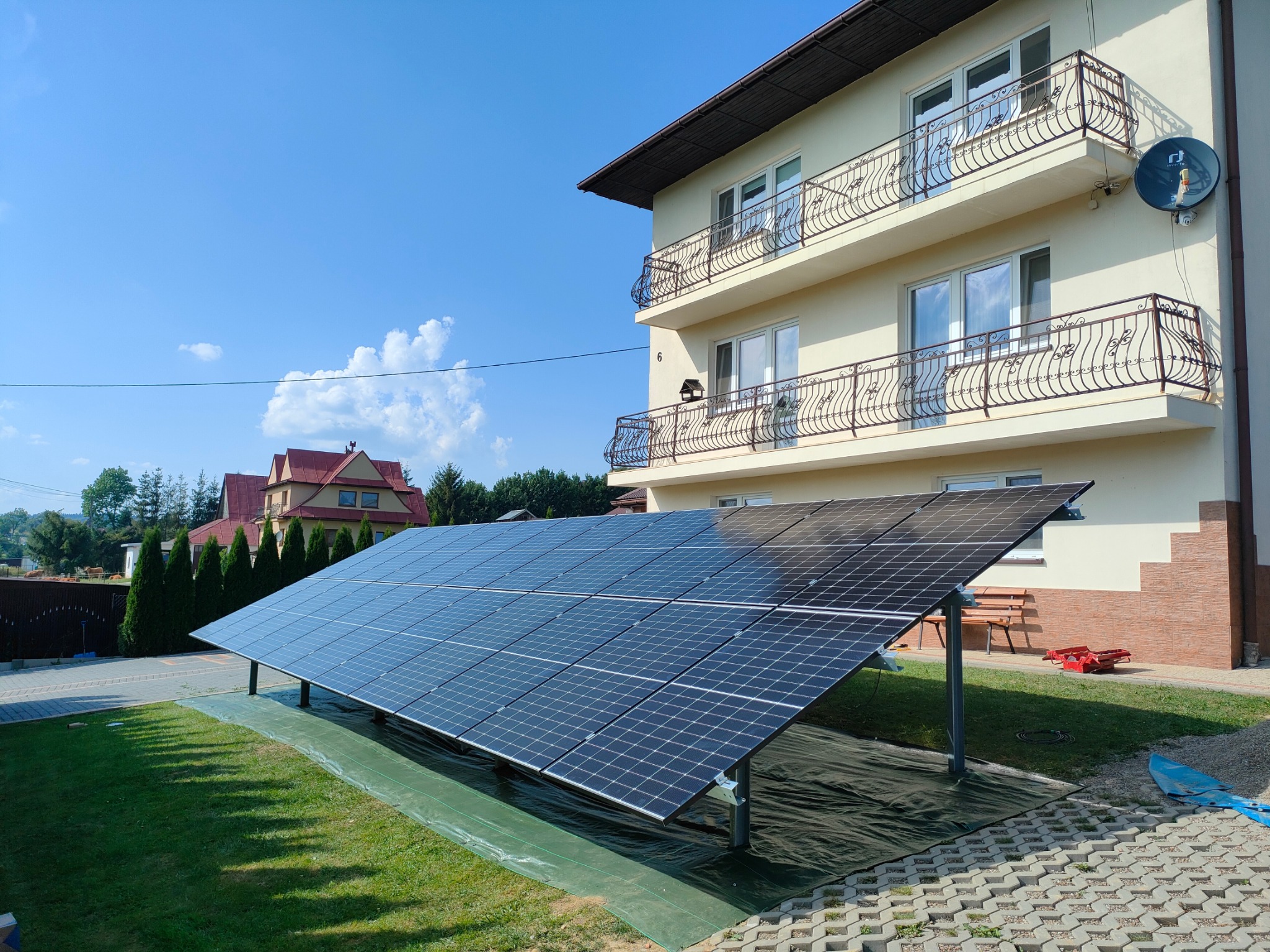 Panel fotowoltaiczny na trawniku przed domem z balkonami. Instalacja na zielonej macie, błękitne niebo w tle. Widok na elewację i ogród.