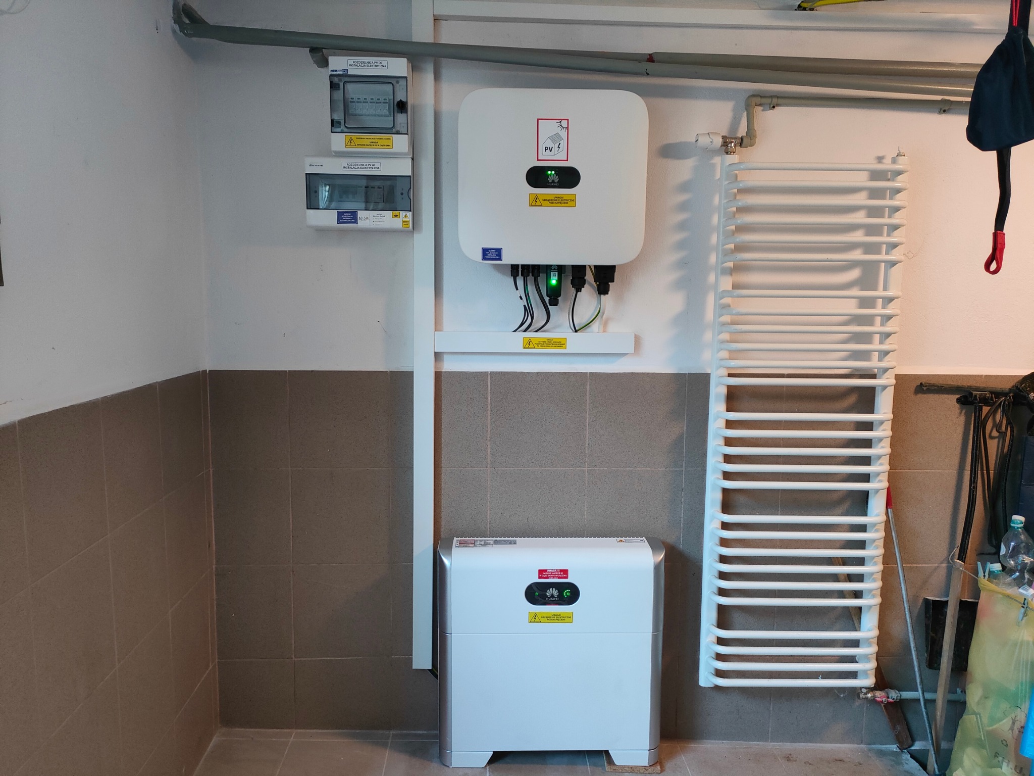 Biały inwerter fotowoltaiczny Huawei i rozdzielnia elektryczna na ścianie w pomieszczeniu w Mochnaczce Niżnej. Widoczny także biały grzejnik drabinkowy.