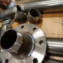 Metalowy kołnierz z przyspawaną rurą, widoczne ślady spawania z charakterystycznym zabarwieniem, w tle inne elementy metalowe i narzędzia na drewnianym stole warsztatowym.