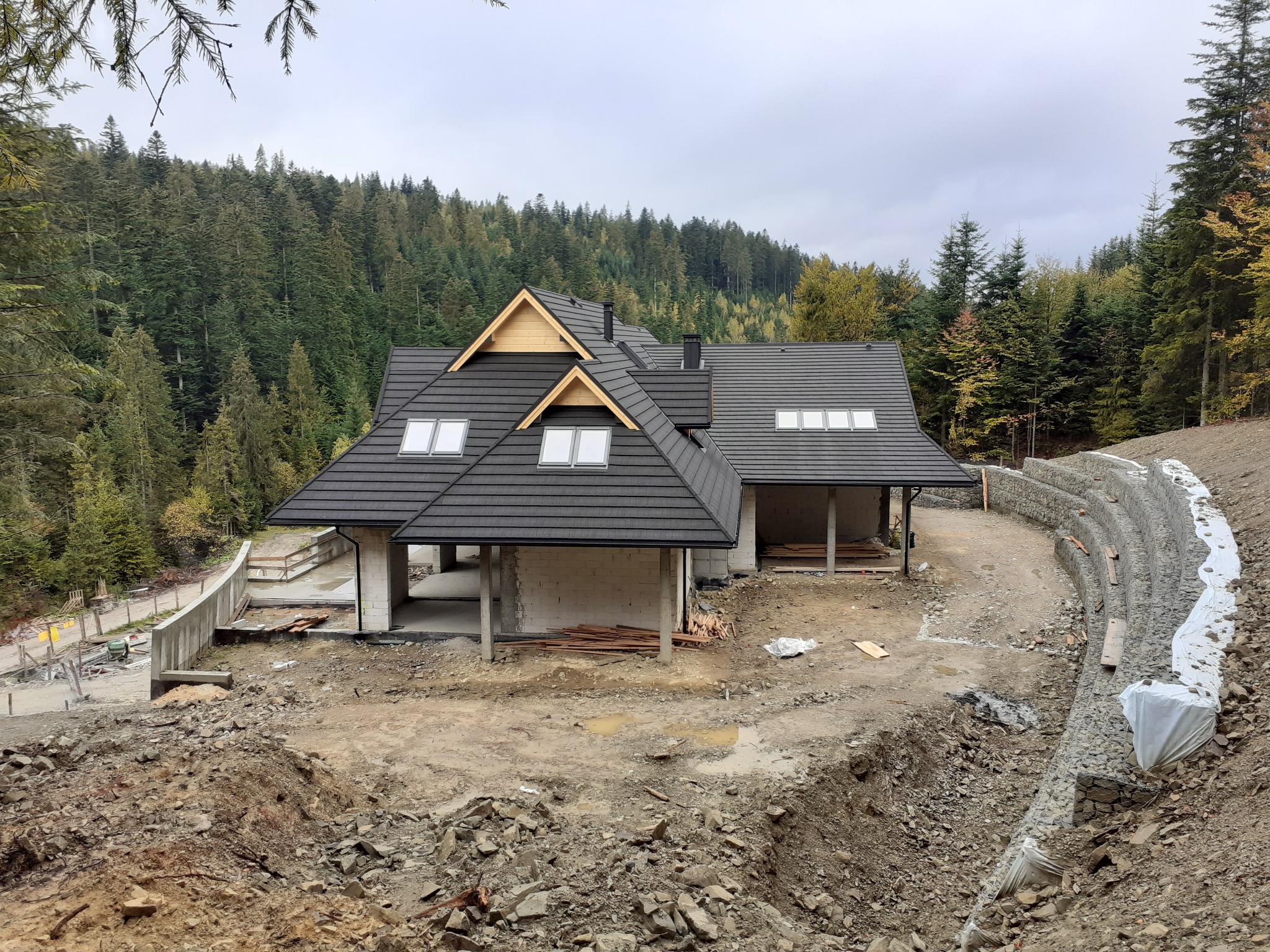 Budynek w stanie surowym otwartym z czarnym dachem, widoczna konstrukcja murowana i teren w trakcie prac ziemnych, w tle las.