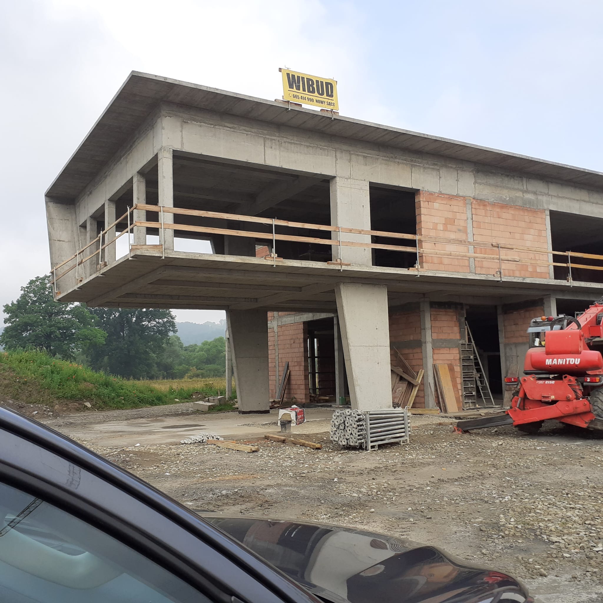 Surowy, dwukondygnacyjny budynek w trakcie budowy z betonu i cegły, wsparty na betonowych filarach. Na dachu widoczna tablica reklamowa firmy Wibud. W tle zieleń drzew i wzgórz, a na pierwszym...