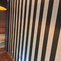 Ściana w pokoju pomalowana w czarno-białe ukośne pasy, zabezpieczona niebieską taśmą malarską przy suficie i ścianie bocznej.