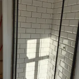 Świeżo położone, białe, prostokątne płytki w kabinie prysznicowej z czarnymi profilami, widoczne otwory na baterię, cień pada na ścianę