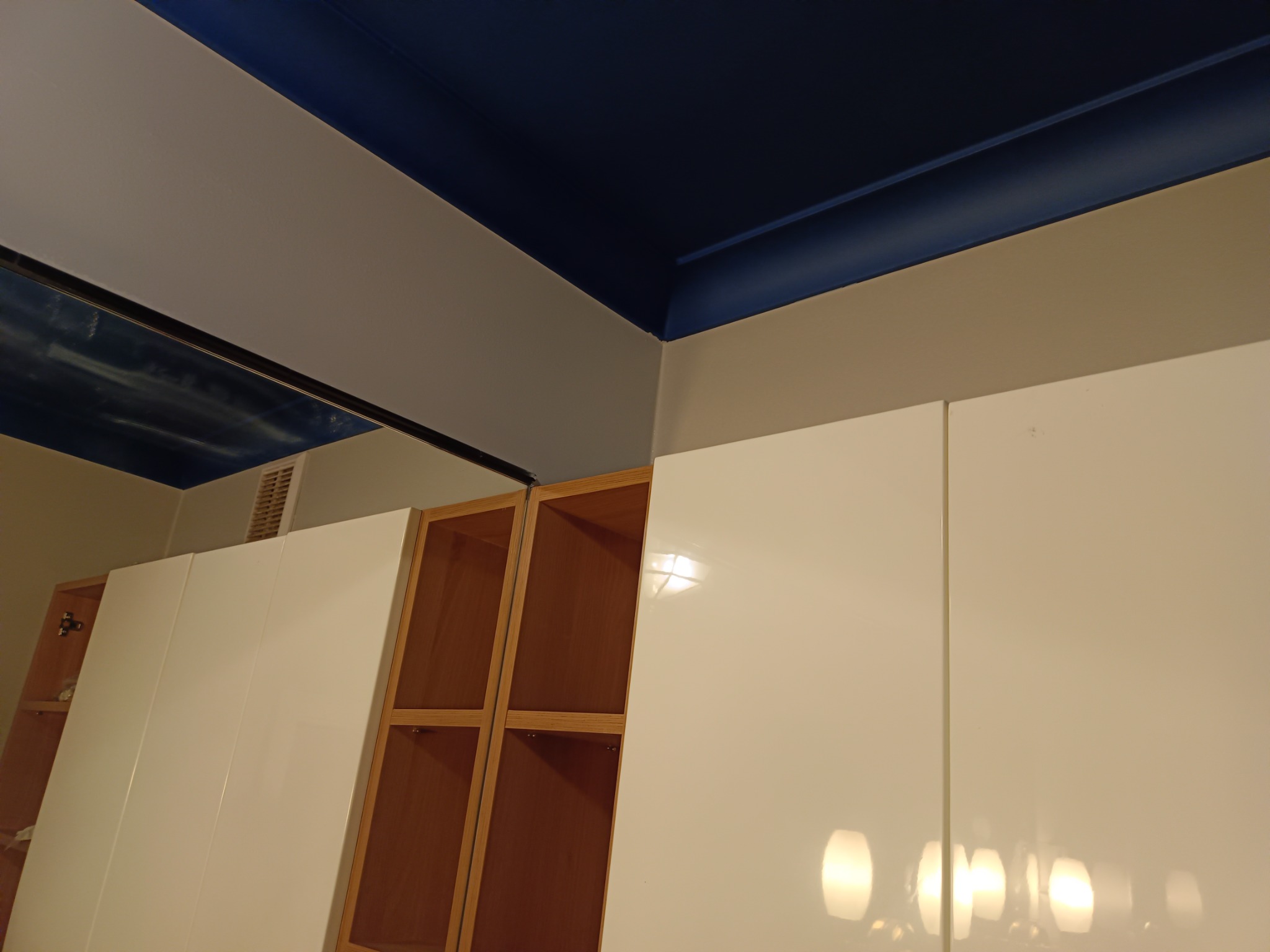 Pomieszczenie z pomalowanym na granatowo sufitem, jasnymi ścianami i białymi szafkami z drewnianymi regałami w tle.