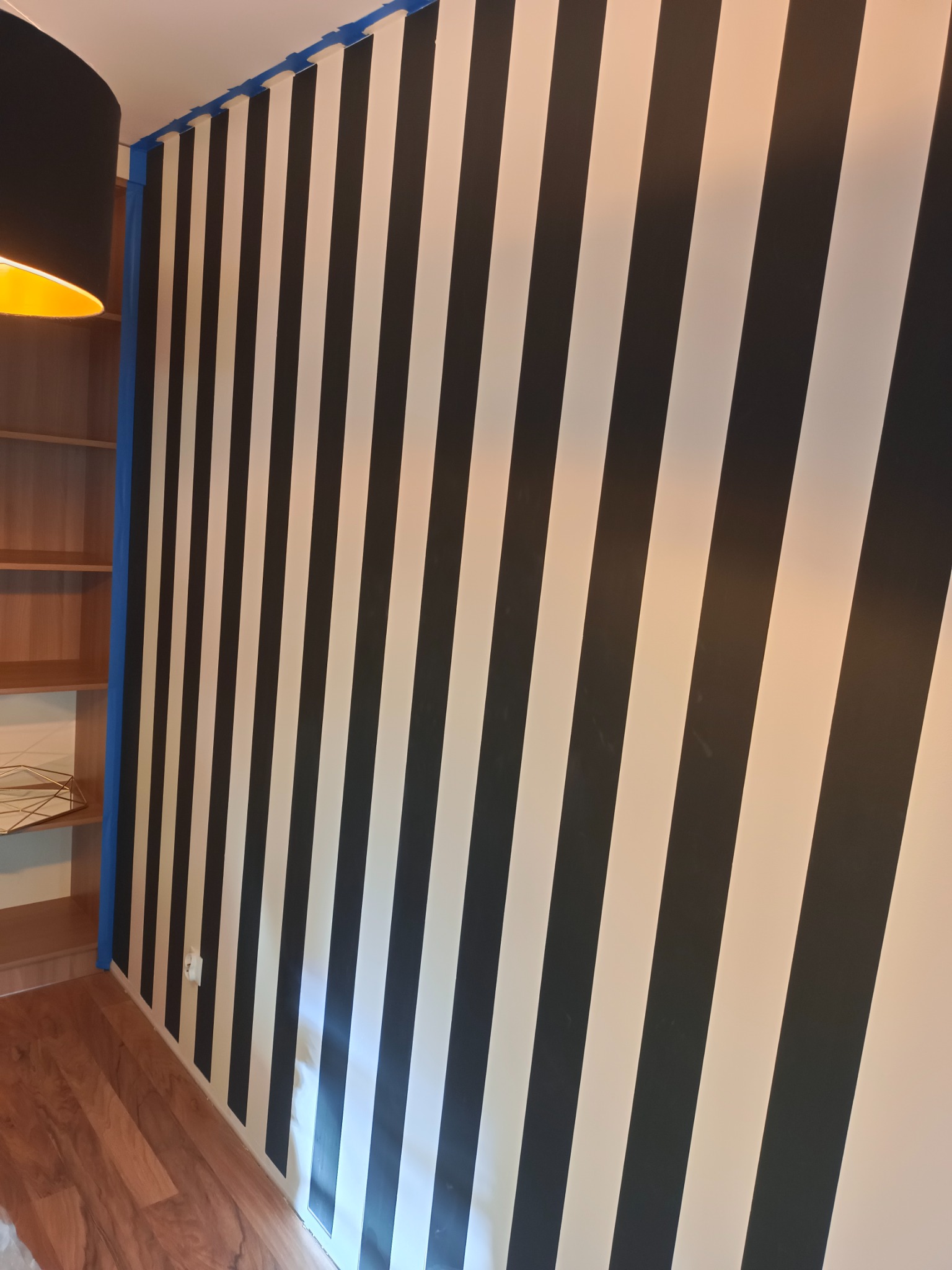 Ściana w pokoju pomalowana w czarno-białe ukośne pasy, zabezpieczona niebieską taśmą malarską przy suficie i ścianie bocznej.
