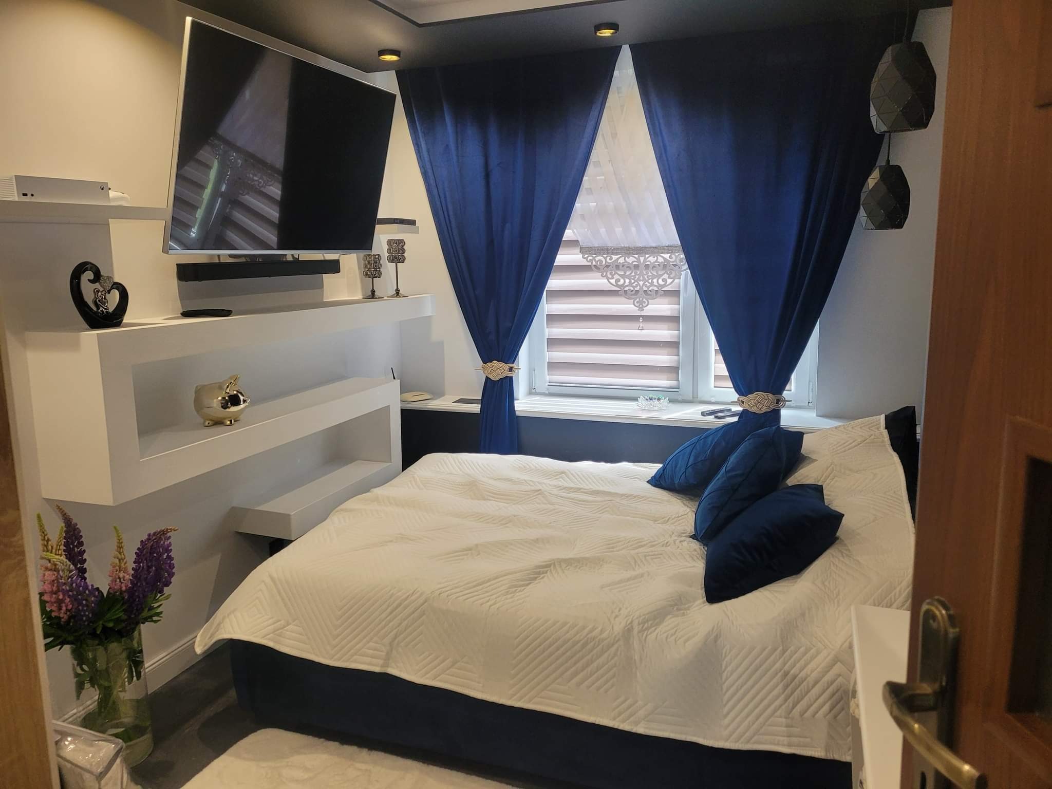 Nowoczesna sypialnia z białymi półkami, telewizorem, dekoracjami, niebieskimi zasłonami i pościelą w geometryczne wzory, z ciemnym sufitem i lampami wiszącymi.