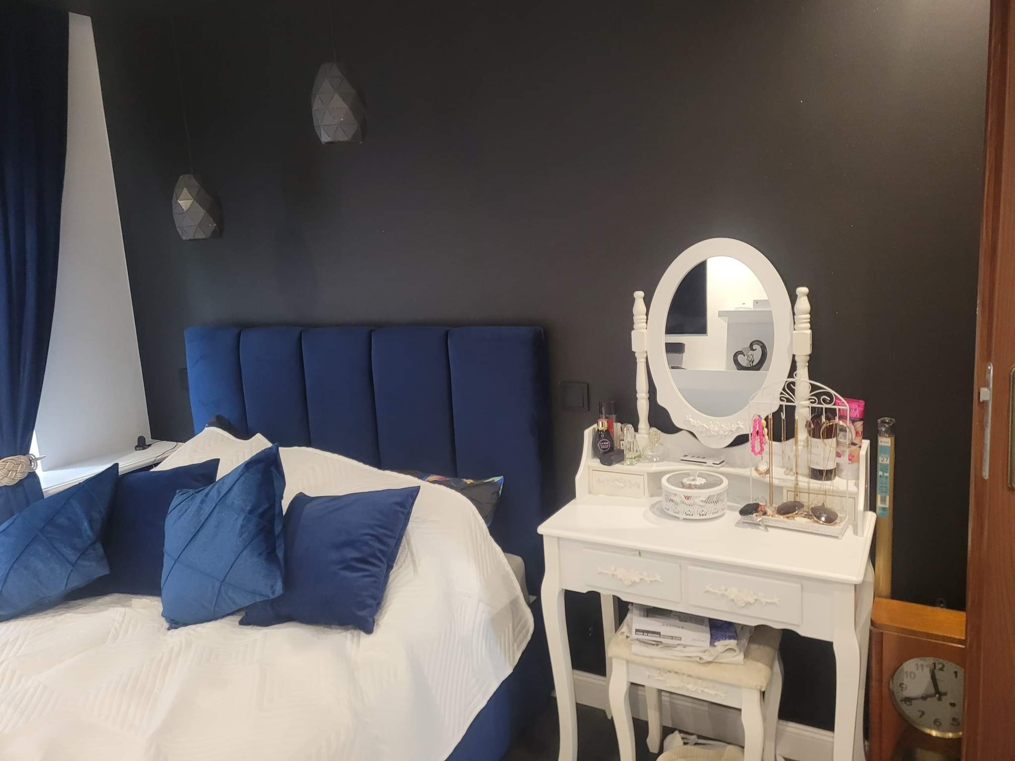 Sypialnia z ciemną ścianą, niebieskim tapicerowanym zagłówkiem, białym łóżkiem z niebieskimi poduszkami i białą toaletką z owalnym lustrem i różnymi akcesoriami.