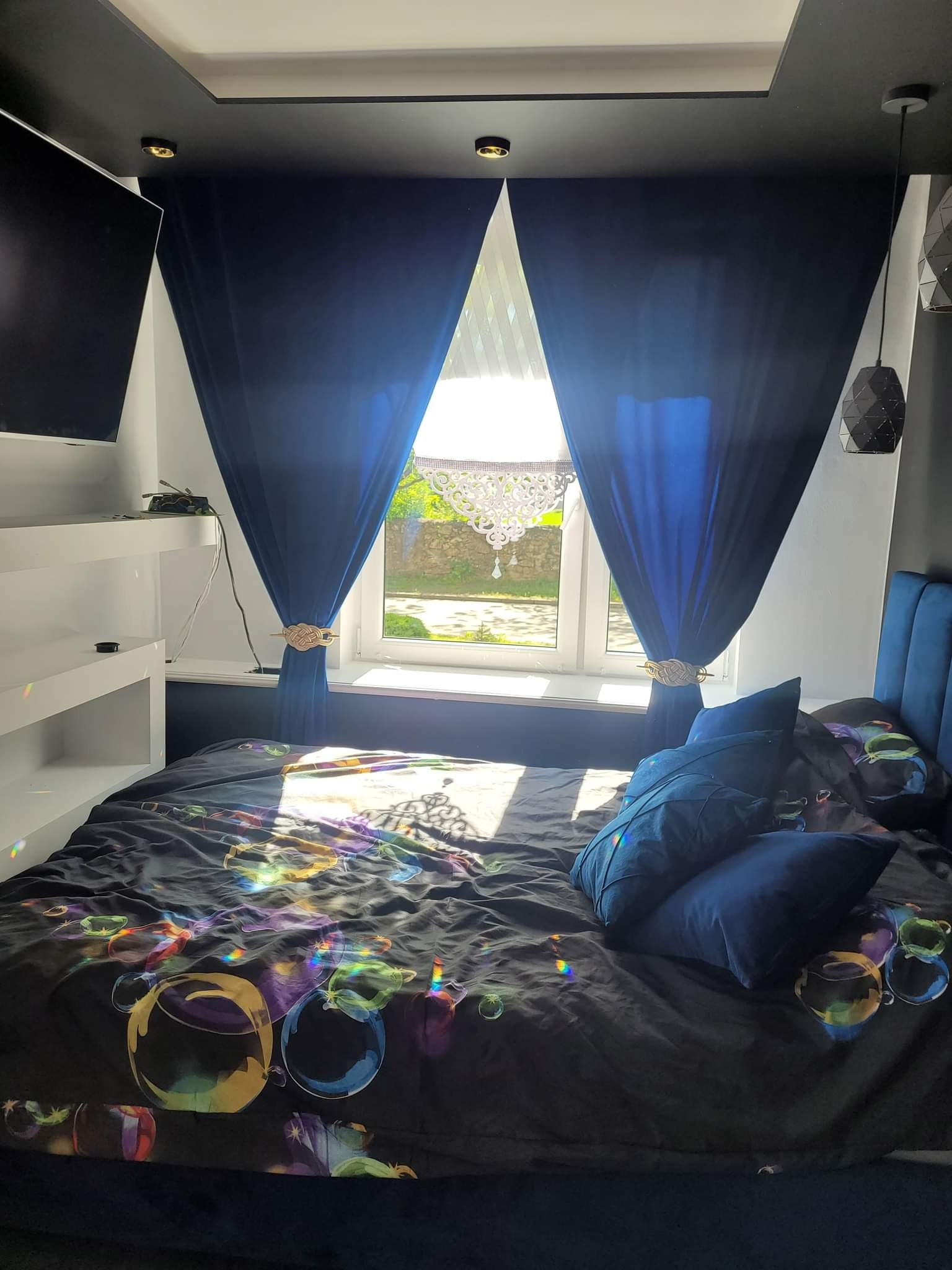 Sypialnia z ciemną, podwieszaną zabudową sufitu, granatowymi zasłonami spiętymi ozdobnymi klamrami, łóżkiem z granatowym zagłówkiem i pościelą w kolorowe bańki mydlane.