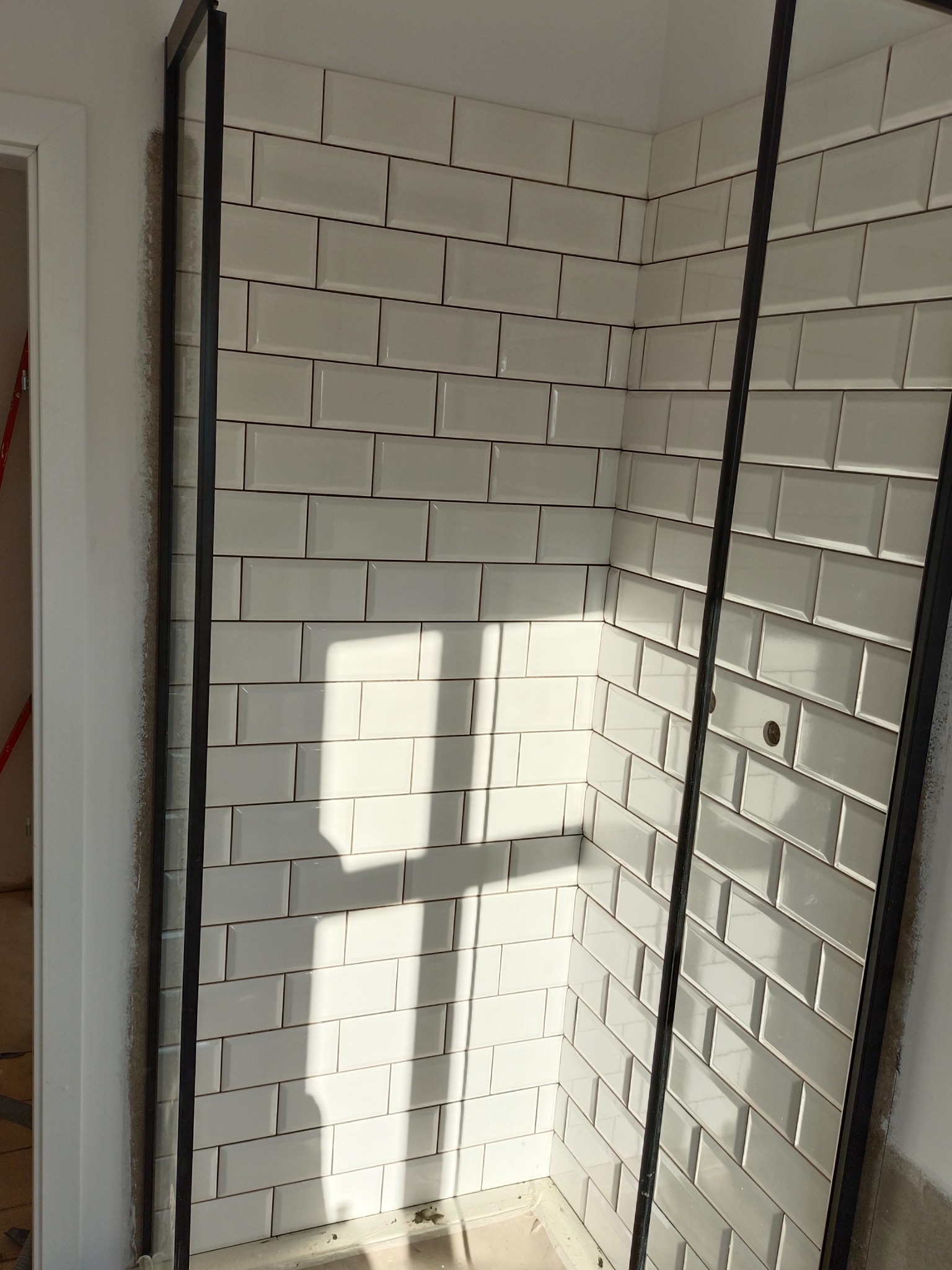 Świeżo położone, białe, prostokątne płytki w kabinie prysznicowej z czarnymi profilami, widoczne otwory na baterię, cień pada na ścianę