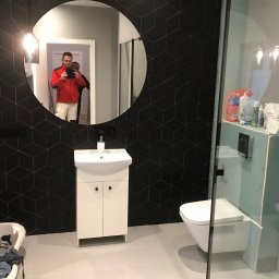 Tomasz Rogaczewski - Nowoczesna łazienka z czarnymi płytkami w geometryczne wzory, okrągłe lustro odbijające osobę robiącą zdjęcie, biała umywalka z szafką i toaleta w trakcie instalacji za szklaną ścianką prysznicową.